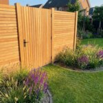 Teakholz Gartentor mit 5 cm Lamellen in einem Teak Zaun hinter einem Beet mit Ziergräsern